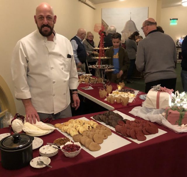 Jamie Muschett stands in a white chef coat smiling. He is behind two tables in an L-shape, which are holding the glorious array of holiday sweets he has made for the event.
