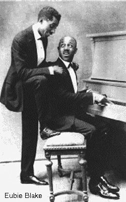 Eubie Blake and Noble Sissle are both in tuxes. Noble sits at the piano, playing, and looking back at Eubie who is standing behind him with hand on Noble's shoulders.