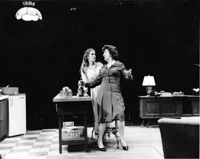 They are standing in the middle of a kitchen set on stage, Maureen in front of the table gesticulating strongly, and Lucy behind her looking on.