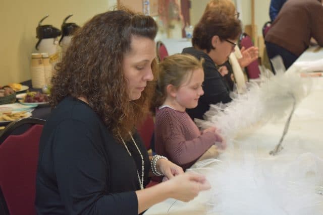 Parents and children are seated around a white table shaping tulle and wire into squirrel tails. Everyone is intensely concentrating on the task at hand, though some are smiling while they work.