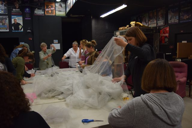 Parents are gathered around a white table with crafting supplies. One mother is holding a long swath of tulle, others are cutting out lengths of various fabrics.