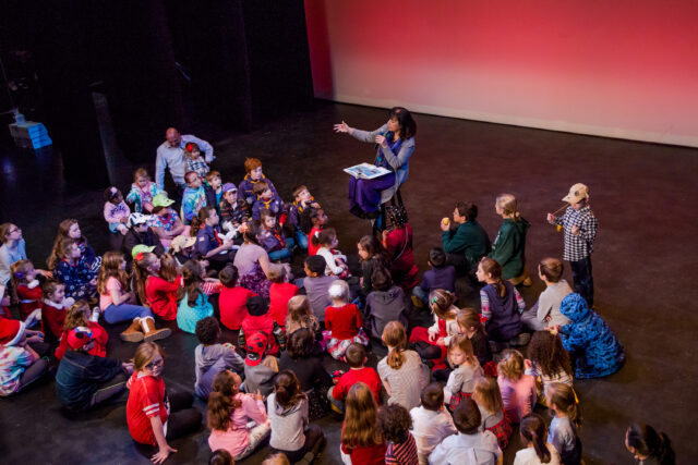 Kate Maguire sits on a stool on stage with a book open in her lap and a microphone. She is reading to a large group of children in front of her, also on stage.