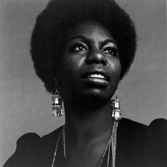 Photo of Nina Simone wearing a dark top with long dangly earrings, looking up and to the left.